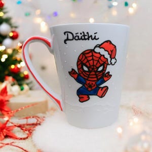 May include: White ceramic mug with a red and blue cartoon image of Spiderman wearing a Santa hat. The mug has a red handle and the name "D&aacute;ith&iacute;" written on the front.