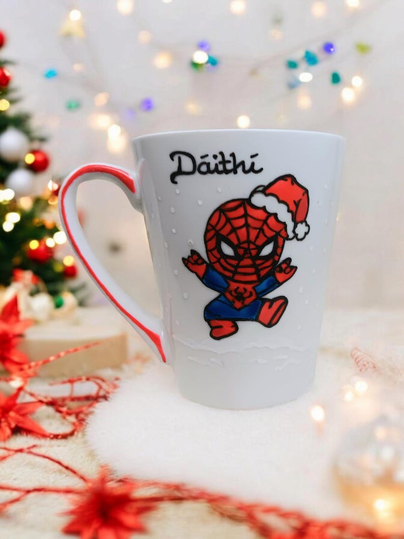 May include: White ceramic mug with a red and blue cartoon image of Spiderman wearing a Santa hat. The mug has a red handle and the name "D&aacute;ith&iacute;" written on the front.