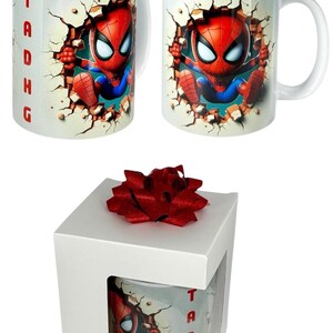 May include: White ceramic mug featuring a cartoon Spider-Man character breaking through a wall. The mug has a white handle and the name "Tadhg" in red letters. The mug is in a white gift box with a red bow.