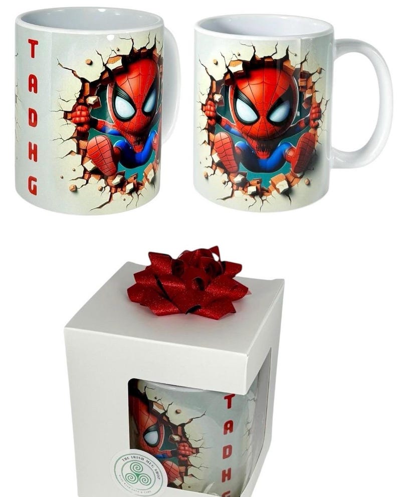 May include: White ceramic mug featuring a cartoon Spider-Man character breaking through a wall. The mug has a white handle and the name "Tadhg" in red letters. The mug is in a white gift box with a red bow.