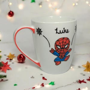 May include: White ceramic mug with a red rim and a cartoon image of Spiderman. The mug has the name "Luke" written on it.