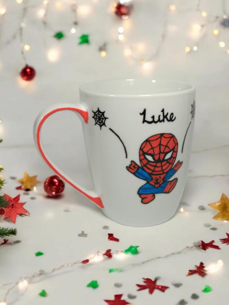 May include: White ceramic mug with a red rim and a cartoon image of Spiderman. The mug has the name "Luke" written on it.