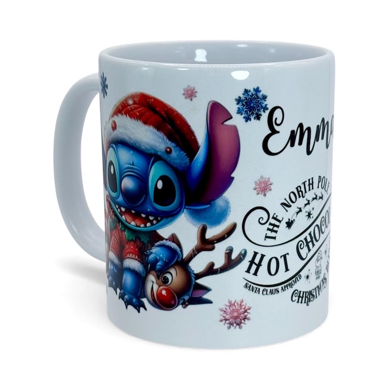 May include: White ceramic mug featuring a cartoon character in a Santa hat and a reindeer. The mug has the text "Emma" and "The North Pole Hot Chocolate Christmas Santa Claus Approved". Decorative snowflakes are also present.