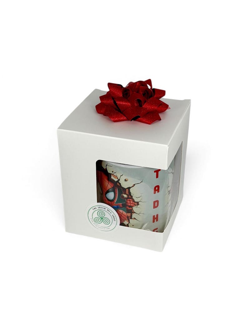 May include: A white gift box with a red bow on top. The box has a window showing a Spider-Man mug. The mug features a graphic of Spider-Man breaking through a wall, with the name "Tadhg" in red.