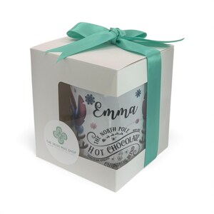 May include: A white gift box tied with a teal ribbon, showcasing a mug with the name "Emma" and the text "The North Pole Hot Chocolate Christmas Blend." The mug features snowflake and reindeer designs.