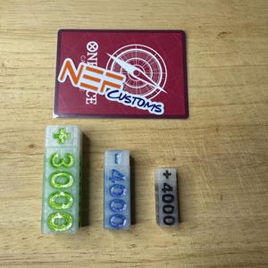 Pode incluir: Três peças de jogo retangulares e translúcidas com números e um sinal de mais, em verde, azul e branco. Um cartão vermelho com texto branco e um desenho de bússola está acima deles. O texto no cartão diz "ONE PIECE CUSTOMS".