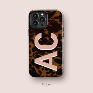 Op de afbeelding: Een rechthoekig smartphonehoesje met een bruin en zwart schildpadpatroon. De letters AC zijn in een groot, lichtroze schreefloos lettertype met een witte omlijning in het midden op de achterkant gedrukt. De uitsparing voor de camera is zichtbaar.