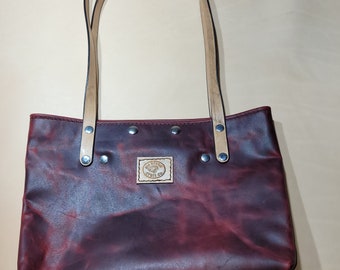 May include: A burgundy leather tote bag with tan leather straps and a small brown leather patch with the text "The Prairie Craft Co."