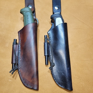 May include: Two leather knife sheaths, one brown and one black, each holding a knife with a green handle. The sheaths have a loop for belt attachment and a small pouch. The knives are designed for outdoor use.