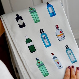 May include: White kitchen towel with a repeating pattern of various bottles of gin. The bottles are illustrated in different colors, including blue, green, and red.