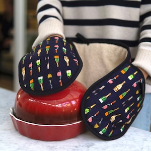 May include: Two oven mitts with a pattern of champagne bottles on a dark blue background. The mitts are stacked on top of a red ceramic baking dish.