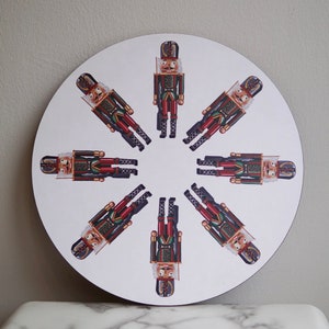 May include: A round white coaster featuring a design of eight nutcracker soldiers in red, green, and gold. The nutcrackers are arranged in a circle around the centre of the coaster.