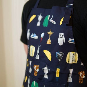 May include: A navy blue apron with a variety of golf-themed illustrations, including golf clubs, golf balls, golf shoes, a golf glove, and a golf umbrella. The apron also features a pocket on the front.