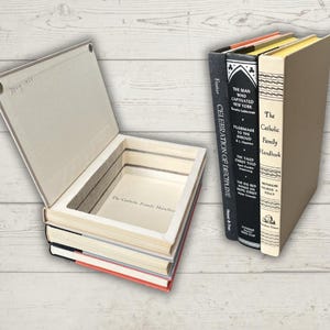 Könnte beinhalten: Ein dekorativer Buchsafe aus recycelten Büchern. Das offene Buch zeigt ein verstecktes Fach. Drei weitere Bücher stehen neben dem offenen Buch. Die Bücher tragen Titel wie "Celebration of Discipline" und "The Catholic Family Handbook".