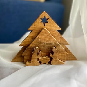 Peut inclure: Une crèche en bois composée d'un fond triangulaire en forme de sapin avec une étoile découpée au sommet. Devant, trois figurines en bois distinctes sont disposées : deux personnages agenouillés face à une mangeoire centrale en bois clair.