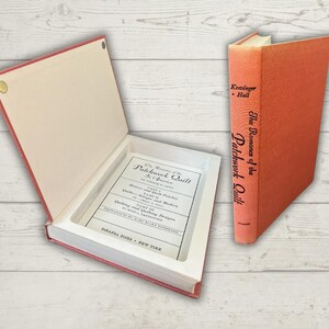 Puede incluir: Un libro abierto, con cubierta roja y un interior empotrado, que revela una página de título. Al lado, un libro cerrado con una cubierta naranja y el título "The Romance of the Patchwork Quilt" se muestra. Los libros están sobre una superficie de grano de madera clara.