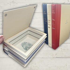 Puede incluir: Una caja fuerte decorativa con forma de libro con un compartimento oculto, exterior color crema y un diseño floral en el interior. La caja fuerte está hecha de libros reutilizados, con un libro azul, rojo y crema visible al lado.