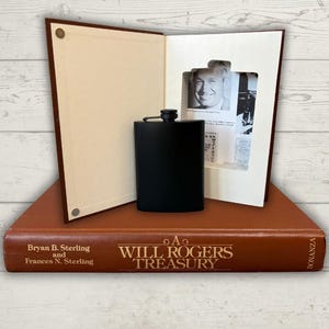 May include: A matte black metal flask stands in front of a hollowed-out book titled A Will Rogers Treasury by Bryan B. Sterling and Frances N. Sterling. The book interior features a black and white portrait of Will Rogers and printed text.