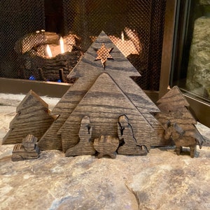 May include: A wooden nativity scene with a star on top of the stable. The figures are cut out of wood and painted in a dark brown color. The scene is set in front of a fireplace.