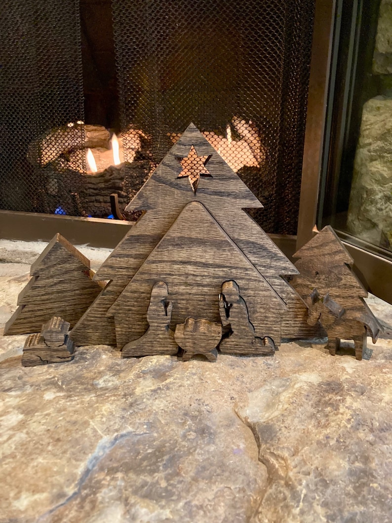May include: A wooden nativity scene with a star on top of the stable. The figures are cut out of wood and painted in a dark brown color. The scene is set in front of a fireplace.