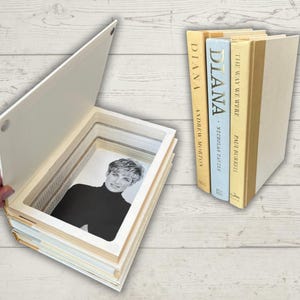 Puede incluir: Una caja decorativa con forma de libro con un retrato de una mujer con un cuello alto negro. La caja está abierta, revelando el retrato dentro de un marco en capas. Al lado, tres libros con los títulos "DIANA" y "THE WAY WE WERE" están de pie.