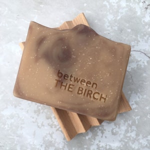 May include: A rectangular bar of soap with a light brown and dark brown marbled design, resting on a wooden soap dish. The words "between THE BIRCH" are embossed on the soap's surface. The soap dish has a slatted design.