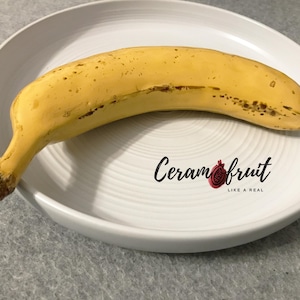 Puede incluir: Un plátano amarillo maduro con manchas marrones descansa sobre un plato de cerámica blanco y estriado. El plato tiene el texto "Ceramfruit" y un logotipo de granada. El plátano está curvado y tiene un tallo marrón.