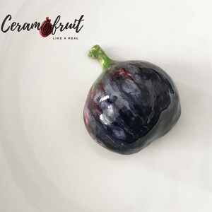 May include: A ceramic fig with a dark purple and blue exterior, a green stem, and a glossy finish. The fig is on a white plate. The text "Ceramfruit LIKE A REAL" is in the upper left corner.