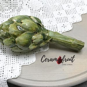 Peut inclure: Une sculpture d'artichaut en céramique dans les tons de vert, avec une longue tige. La sculpture est posée sur une surface en bois gris clair, avec le texte "Ceramfruit" et un logo de grenade. Un napperon en dentelle blanche est en arrière-plan.