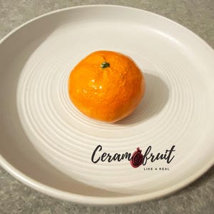 Può includere: Un frutto arancia in ceramica su un piatto bianco. Il frutto è una replica realistica di una vera arancia. Il testo "Ceram fruit like a real" è stampato sul piatto.