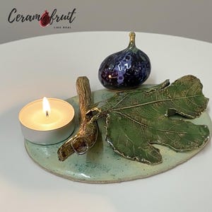 Op de afbeelding: Een keramische kandelaar met een vijgenthema met een brandende theelichtkaars. De houder heeft een groen vijgenblad, een donkerblauwe vijg en een tak. De basis is lichtgroen en de tekst "Ceramfruit LIKE REAL" is zichtbaar.