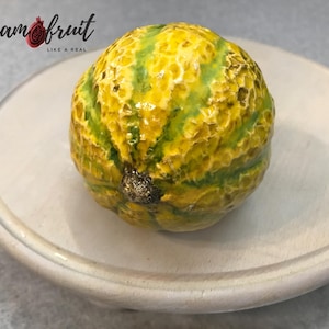 May include: A ceramic melon with a yellow and green rind. The melon is sitting on a white ceramic plate.