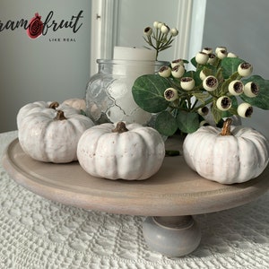 May include: White ceramic pumpkins on a wooden tiered stand with a gray base. The stand is decorated with a sprig of white berries and green leaves. The text "Ceram fruit LIKE REAL" is visible in the background.