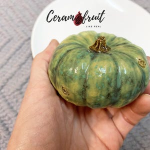 Peut inclure: Une citrouille en céramique émaillée vert et jaune, tenue dans une main. La citrouille a une surface texturée et une tige marron. Le texte "Ceramfruit LIKE REAL" est au-dessus.