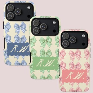 May include: Three phone cases with bow patterns in blue, green, and pink. Each case has a cream background and a different coloured bow pattern. Each case has a rectangular label with the initials "R.W." in a matching colour.