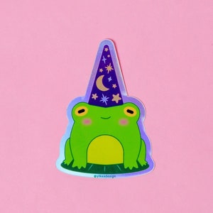 May include: A green frog wearing a purple and blue wizard hat with stars and a crescent moon. The frog is sitting on a green lily pad. The sticker has a holographic finish.