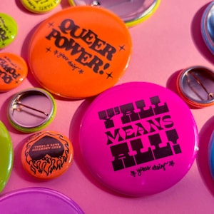 May include: A collection of round pin-back badges in vibrant colours including neon pink, orange, yellow, and lime green. The badges feature black text such as QUEER POWER!, Y'ALL MEANS ALL!, and KEEP ABORTION SAFE & LEGAL, along with star and flame graphics.