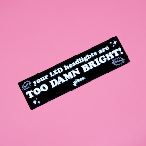 May include: A black rectangular sticker with white text that reads "your LED headlights are TOO DAMN BRIGHT!" with the word "yikes." below. The sticker is set against a pink background.