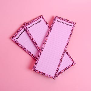 May include: Pink notepads with lined pages and a leopard print border. The text "Don't Forget!" is printed at the top of each notepad. The notepads are arranged on a pink background.