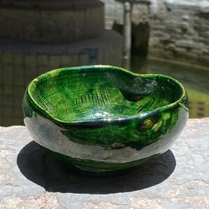 Puede incluir: Un pequeño cuenco de cerámica formado a mano con un esmalte brillante de color verde esmeralda intenso. El cuenco presenta un borde de forma orgánica irregular y marcas de textura verticales visibles en las paredes interiores, con una base redondeada.