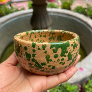 Puede incluir: Un cuenco pequeño y redondo de arcilla color terracota con un patrón moteado de salpicaduras de pintura verde oscuro en su superficie exterior. El cuenco tiene un borde grueso sin esmaltar y una apariencia porosa y texturizada de barro.