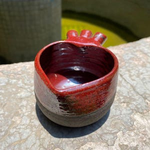 Puede incluir: Un pequeño cuenco de cerámica con un interior y exterior superior esmaltados en rojo brillante, que transiciona hacia una base de color gris carbón mate. El borde presenta una protuberancia distintiva de tres puntas en un lado.