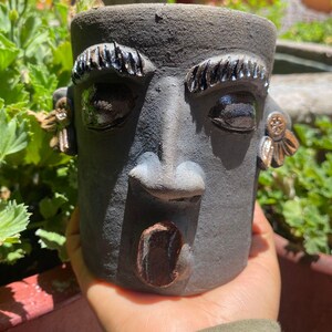 Puede incluir: Una maceta cilíndrica de cerámica gris oscuro con forma de rostro humano y acabado mate texturizado. Presenta cejas negras brillantes en relieve, ojos almendrados, una nariz prominente, una boca abierta y adornos en forma de orejas a los lados.