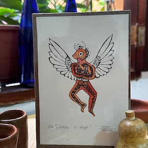May include: A hand-drawn illustration of a winged figure with a red and orange body, black eyes, and a white halo. The figure is standing on one leg and has its arms outstretched. The text below the figure reads "P/a "Diablo o Angel?" Ramirez Ilustraciones".