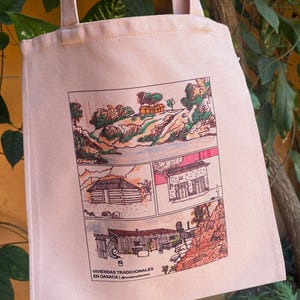 Puede incluir: Un bolso tote rosa claro con una ilustración de varios paneles de casas y paisajes tradicionales. La obra de arte está en un estilo de cómic con acentos verdes, marrones y rojos. El texto "VIVIENDAS TRADICIONALES EN OAXACA" está impreso en la parte inferior.