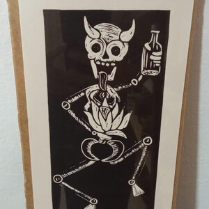May include: Black and white linocut print of a skeletal figure with horns holding a bottle. The figure has a flower-like design in its chest. The print is in a wooden frame.