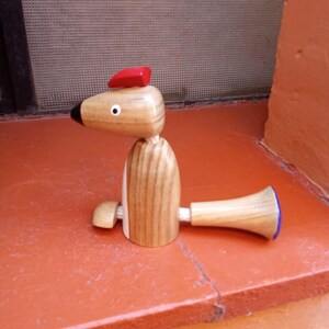 Puede incluir: Un juguete de madera en forma de animal con un sombrero rojo y una cola a rayas azul y blanca. El juguete está hecho de madera natural y tiene un diseño simple y caprichoso.