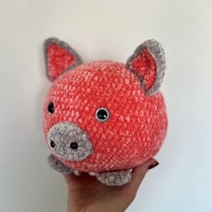 Puede incluir: Un juguete de cerdo suave y redondo en tonos coral y gris. El cerdo tiene un aspecto texturizado y aterciopelado, con detalles grises en las orejas, el hocico y las patas. Los ojos son negros con purpurina plateada.