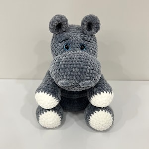 Pode incluir: Um hipopótamo de pelúcia cinza crochetado com detalhes brancos nos pés. O hipopótamo tem olhos azuis.