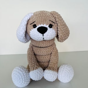 Peut inclure: Un chien en peluche crocheté avec une fourrure brune et des accents blancs. Le chien est assis les pattes croisées et a une expression triste sur son visage.